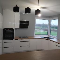 Usługi Stolarskie Marcin Biesiadecki - Nowoczesna kuchnia z białymi szafkami na wymiar, drewnianym blatem, czarnym piekarnikiem i płytą grzewczą, oświetlona trzema czarnymi lampami wiszącymi z żarówkami Edisona, z widokiem na zewnątrz...