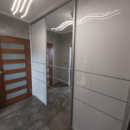 Usługi Stolarskie Marcin Biesiadecki - Biała szafa przesuwna z lustrem, z poziomymi podziałami, w nowoczesnym wnętrzu z drewnianymi drzwiami i szarą podłogą z płytek.