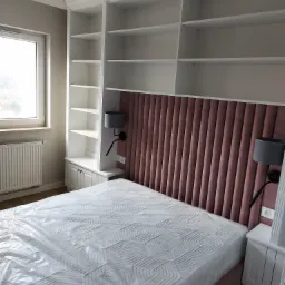 Biała zabudowa meblowa z półkami nad łóżkiem z tapicerowanym, pionowo żebrowanym zagłówkiem w odcieniu pudrowego różu, uzupełniona o szafki nocne i lampki ścienne z regulowanym ramieniem.