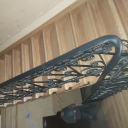 Usługi Stolarskie Marcin Biesiadecki - Kute, czarne balustrady schodowe z dekoracyjnymi elementami spiralnymi, widoczne od dołu, z drewnianymi stopniami w tle i fragmentem osoby w dolnej części kadru.