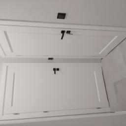 Usługi Stolarskie Marcin Biesiadecki - Dwa białe, prostokątne skrzydła drzwiowe z czarnymi klamkami i zamkiem, zamontowane w suficie, widoczne z dołu, z fragmentem szarej ściany w tle.