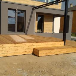 Usługi Stolarskie Marcin Biesiadecki - Drewniany taras z dwoma stopniami, wykonany z jasnych desek, przylegający do nowoczesnego domu z szarą elewacją i czarnymi ramami okiennymi, widoczny mężczyzna w roboczym ubraniu.
