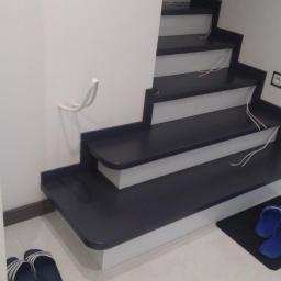 Usługi Stolarskie Marcin Biesiadecki - Nowoczesne schody wewnętrzne z ciemnymi stopniami i białymi podstopnicami, widoczne przewody elektryczne na stopniach, białe ściany, kapcie na wycieraczce u dołu schodów.