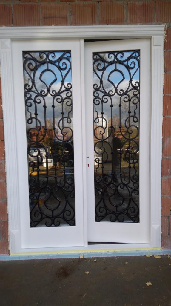 Białe, dwuskrzydłowe drzwi z ozdobnymi, metalowymi kratami w szybach, zamontowane w surowym murze z czerwonej cegły.