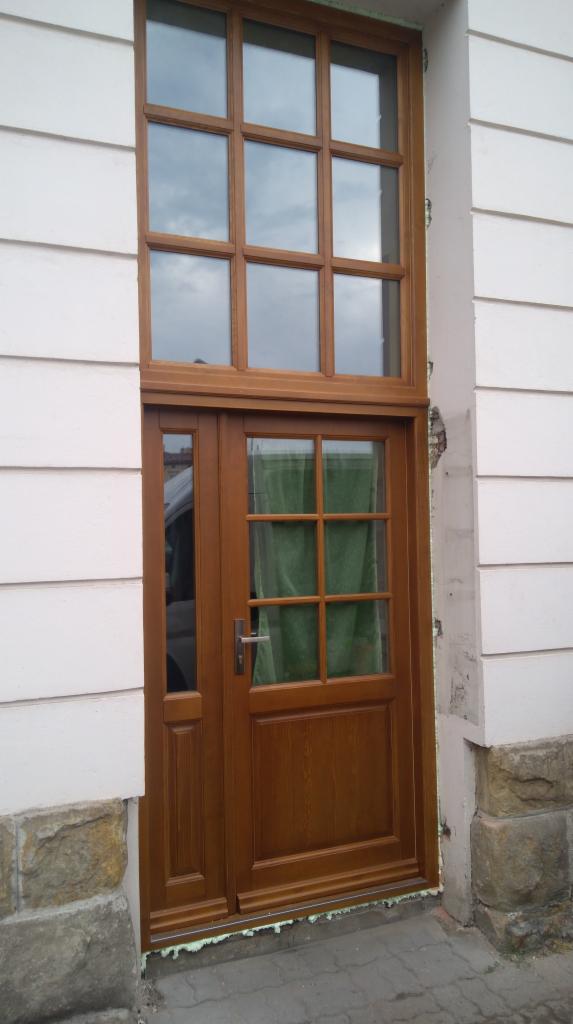Brązowe drzwi wejściowe z szybami, osadzone w białej ścianie z widocznym uszczelnieniem pianką montażową u dołu.