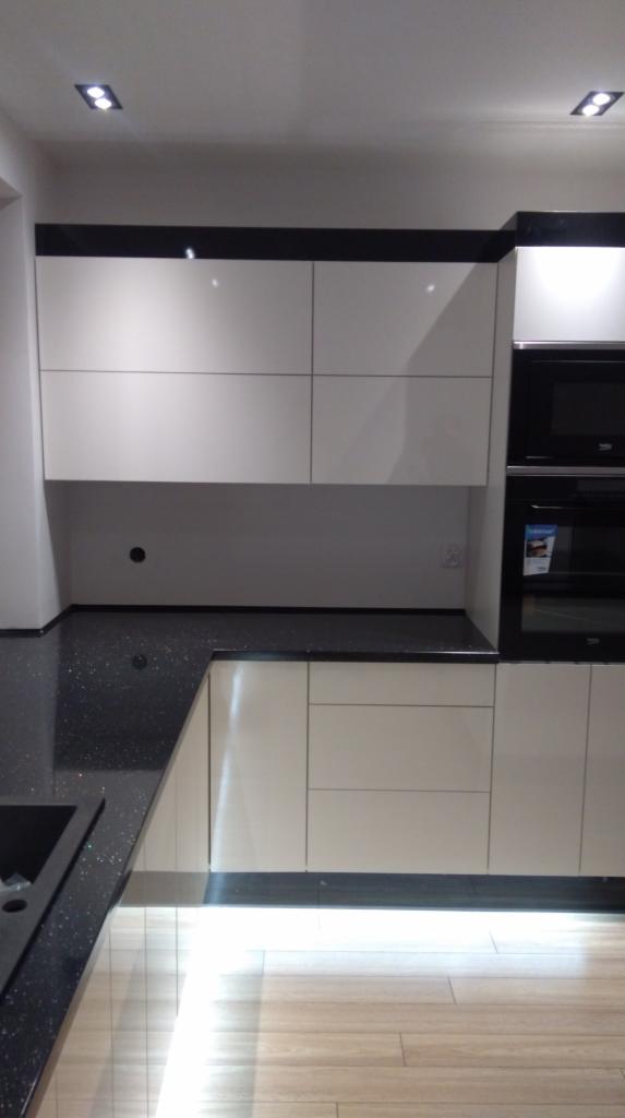 Nowoczesna kuchnia z białymi, lakierowanymi frontami szafek, czarnym blatem z efektem brokatu i oświetleniem LED pod szafkami dolnymi, na podłodze parkiet w jasnym odcieniu drewna.