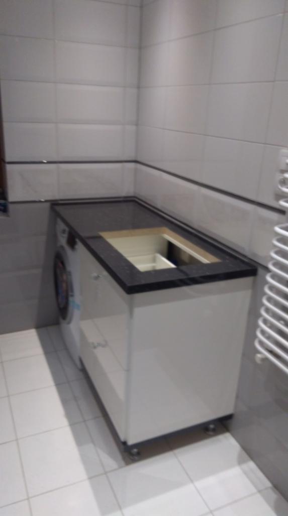 Biała szafka łazienkowa z czarnym blatem, z otworem na umywalkę, stojąca obok pralki w narożniku wyłożonym białymi płytkami.