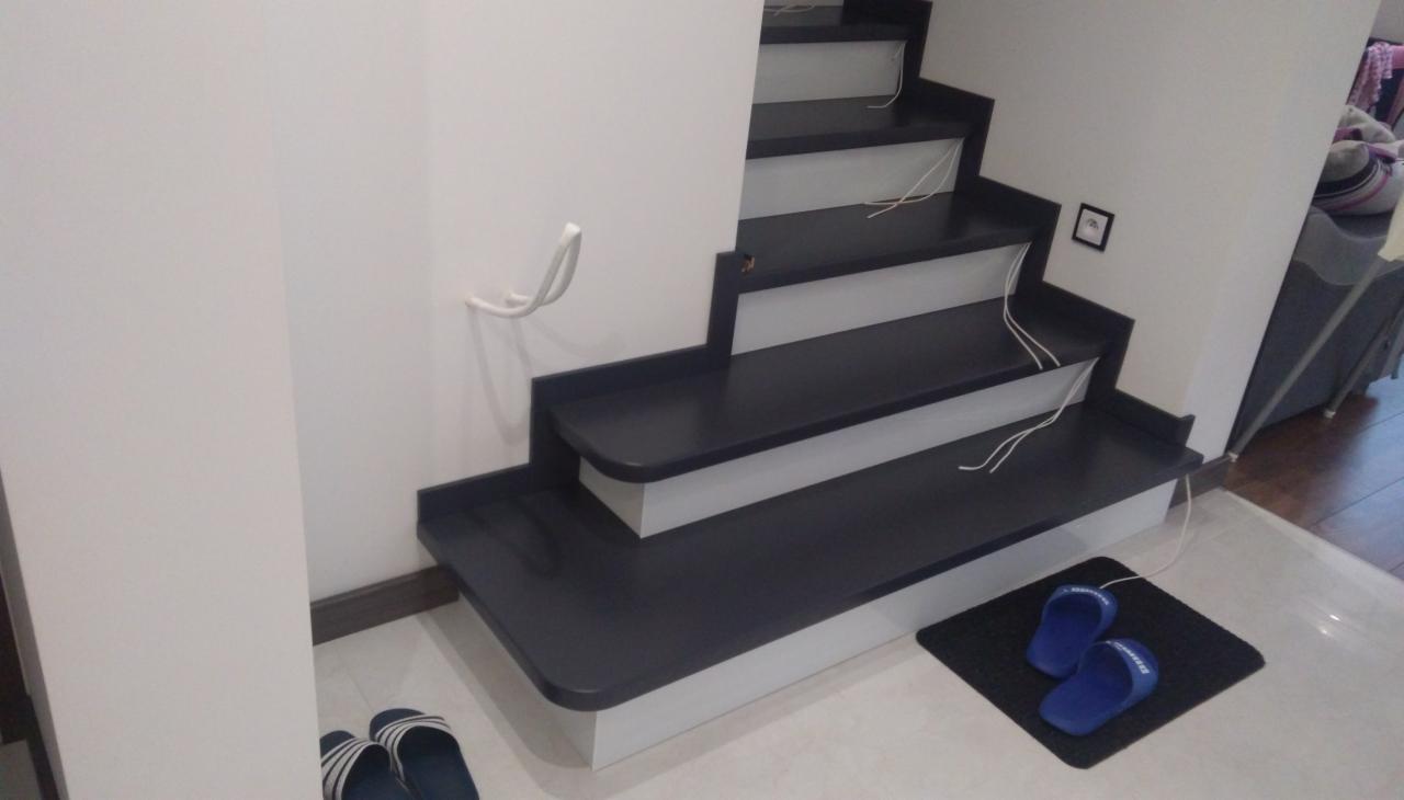 Nowoczesne schody wewnętrzne z ciemnymi stopniami i białymi podstopnicami, widoczne przewody elektryczne na stopniach, białe ściany, kapcie na wycieraczce u dołu schodów.