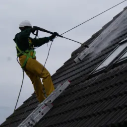 Pracownik w kasku i uprzęży, stojący na drabinie opierającej się o stromy dach z oknami, czyści dach strumieniem wody pod ciśnieniem.