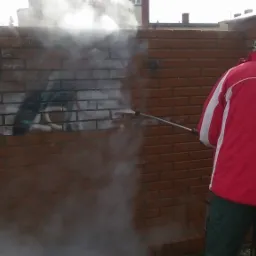 Mężczyzna w czerwonej kurtce czyści ceglany mur z graffiti za pomocą myjki ciśnieniowej, strumień pary wodnej usuwa farbę, widoczne budynki w tle.
