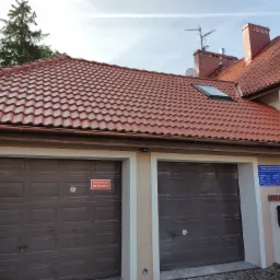 Dom z nową czerwoną dachówką, widoczny komin, antena satelitarna, okno dachowe, dwustanowiskowy garaż z brązowymi bramami segmentowymi, tabliczka informacyjna na ścianie.