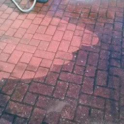 Częściowo oczyszczona z ciemnego nalotu kostka brukowa w kolorze czerwonym, spryskiwana wodą pod ciśnieniem z węża przez osobę w zielonych spodniach roboczych.