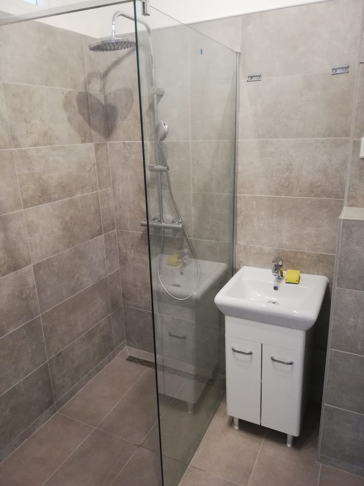 Wykończona łazienka z kabiną prysznicową typu walk-in, szare płytki na ścianach i podłodze, biała szafka z umywalką i żółtą gąbką obok baterii.