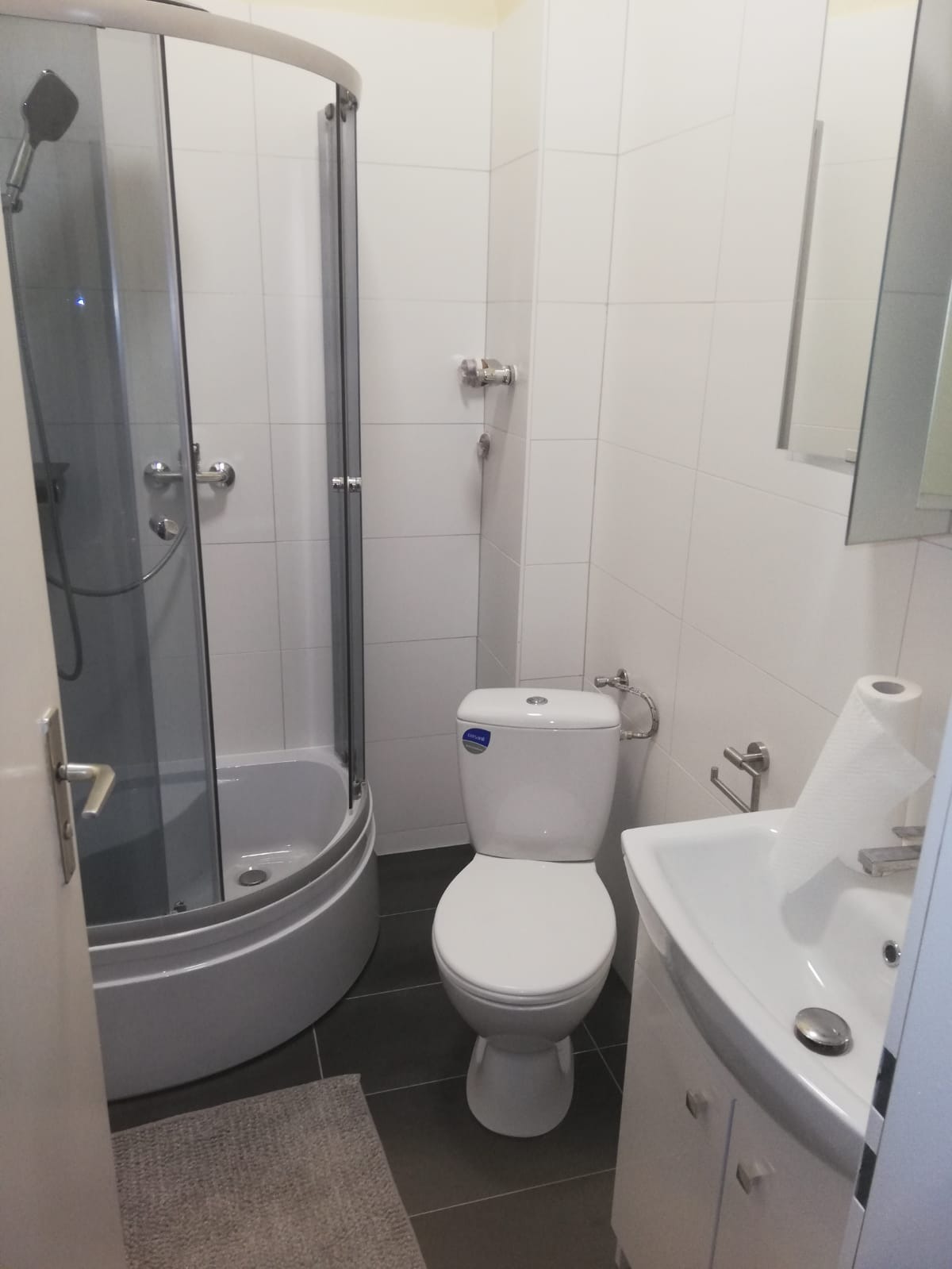 Nowoczesna łazienka z kabiną prysznicową z zaokrąglonym frontem, białą toaletą kompaktową i umywalką z szafką, wykończona białymi płytkami i ciemnoszarą podłogą.