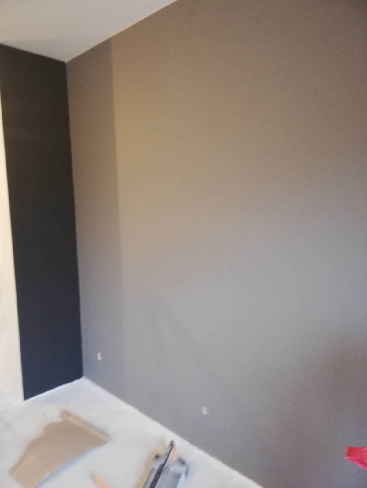 Świeżo pomalowane ściany w odcieniach szarości i grafitu, widoczne narzędzia malarskie i fragment podłogi zabezpieczonej kartonem.
