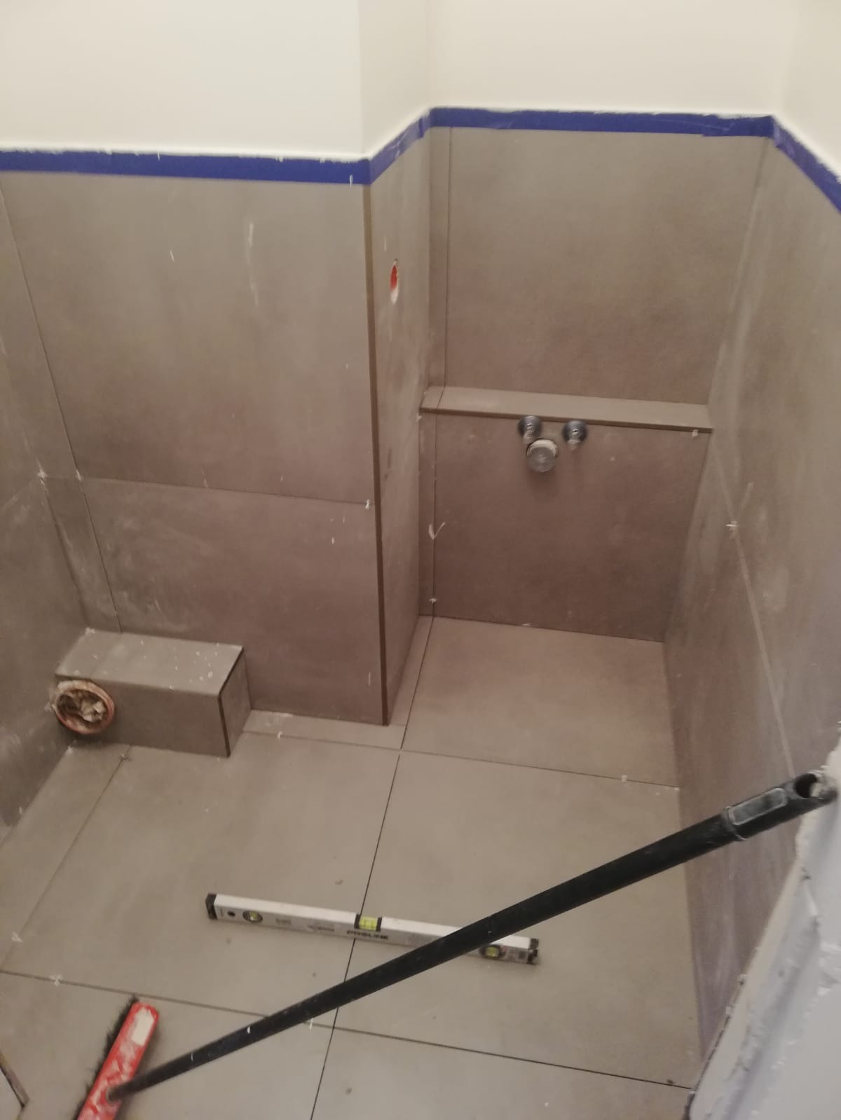 Wnętrze łazienki w trakcie kładzenia płytek, widoczna poziomica, fragment szczotki i wystające rury instalacji wodnej.