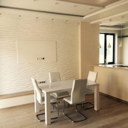 Dobrabud sp. z o.o. - Biała, nowoczesna jadalnia z dekoracyjną ścianą 3D, stołem i krzesłami w odcieniach bieli i beżu. Widok na kuchnię z wiszącymi lampami.