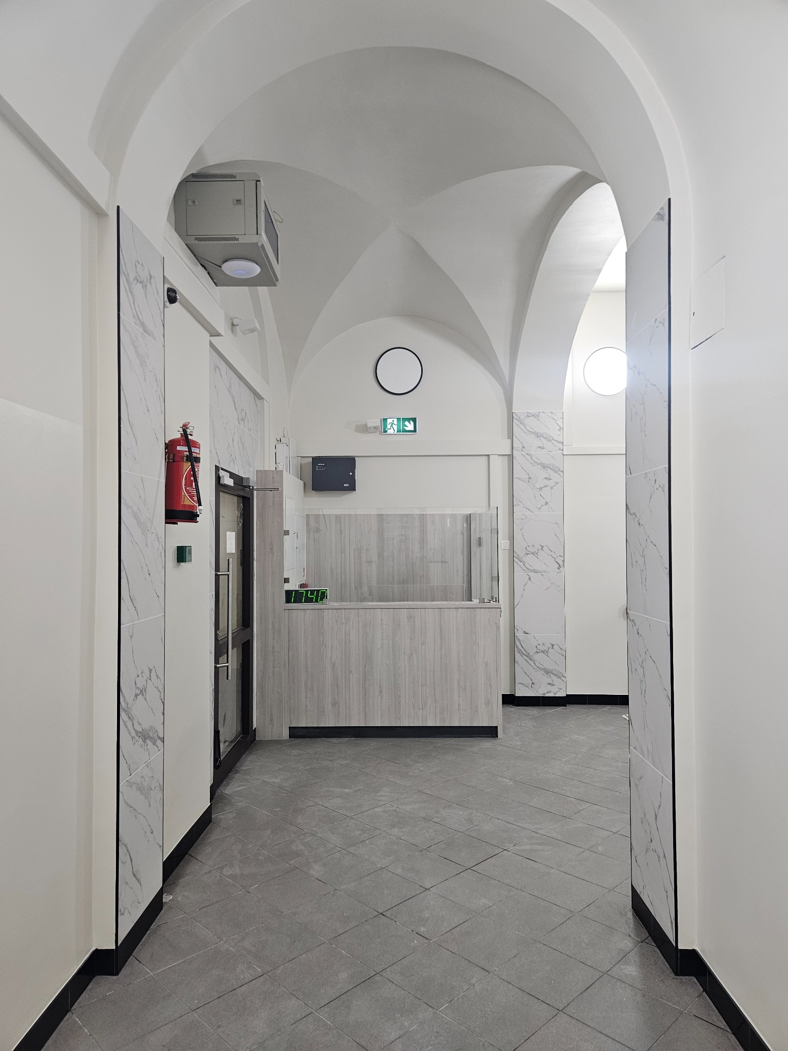 Hall w biurze po remoncie: białe ściany, szara podłoga w płytkach, marmurowe panele i sklepione sufity. Widoczny element recepcji z blatem.