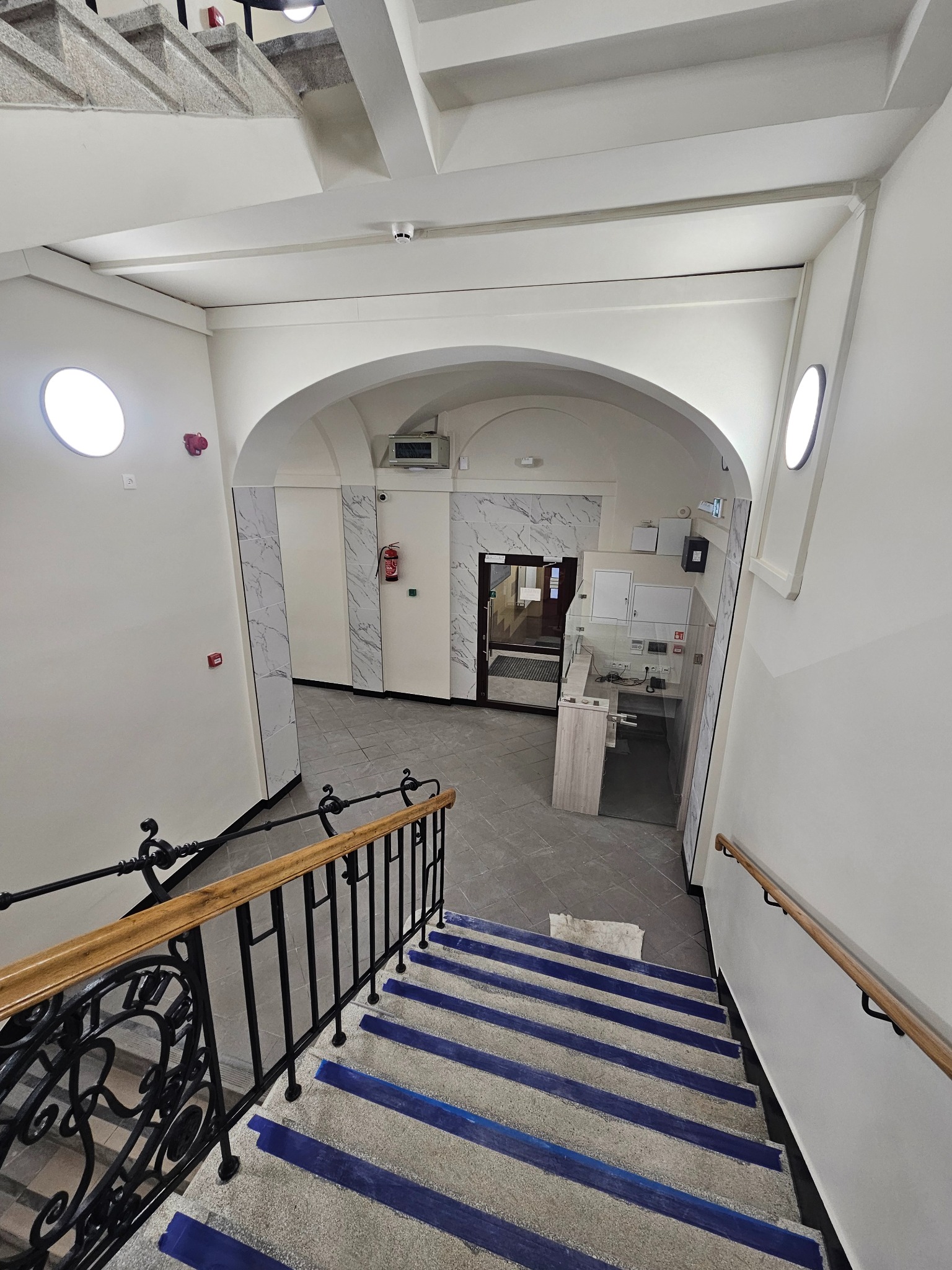 Klatka schodowa w budynku biurowym po remoncie: białe ściany, szare płytki, schody z niebieskimi akcentami, metalowa balustrada, recepcja w tle.