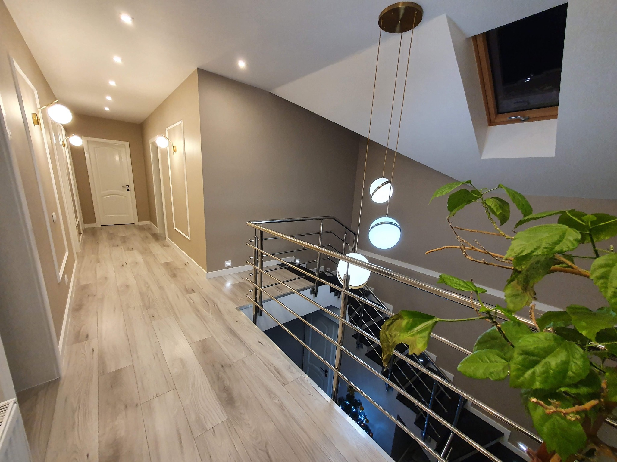 Korytarz z drewnianą podłogą i nowoczesnym oświetleniem w Warszawie. Widoczne schody z metalową barierką i roślina doniczkowa, dodające wnętrzu przytulności.