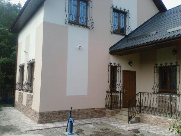 Dwukondygnacyjny dom z jasną elewacją w odcieniach beżu i kremu, z dekoracyjnymi, kutymi kratami w oknach i na balkonie oraz z podmurówką imitującą kamień. Widoczna studnia na pierwszym planie.