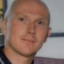 Portret mężczyzny z ogoloną głową w niebieskiej koszuli z białymi paskami. Uśmiechnięta twarz, jasna karnacja, widoczne zmarszczki mimiczne.