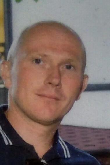 Portret mężczyzny z ogoloną głową w niebieskiej koszuli z białymi paskami. Uśmiechnięta twarz, jasna karnacja, widoczne zmarszczki mimiczne.