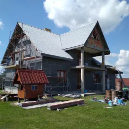 Dom jednorodzinny w trakcie ocieplania elewacji zewnętrznej styropianem, widoczne rusztowania, materiały budowlane i narzędzia na trawniku, na pierwszym planie domek dla dzieci.