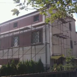 Dwupiętrowy dom w trakcie prac elewacyjnych, z widocznymi rusztowaniami i częściowo pomalowaną fasadą w odcieniach bieli i bordo. Widok zza ogrodzenia.