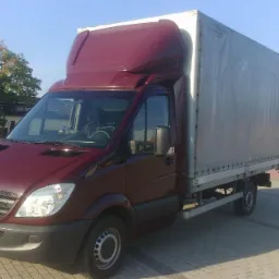 Czerwony bus Mercedes-Benz z plandeką na parkingu w Śmiglu, widok z przodu i boku, z budynkami w tle.