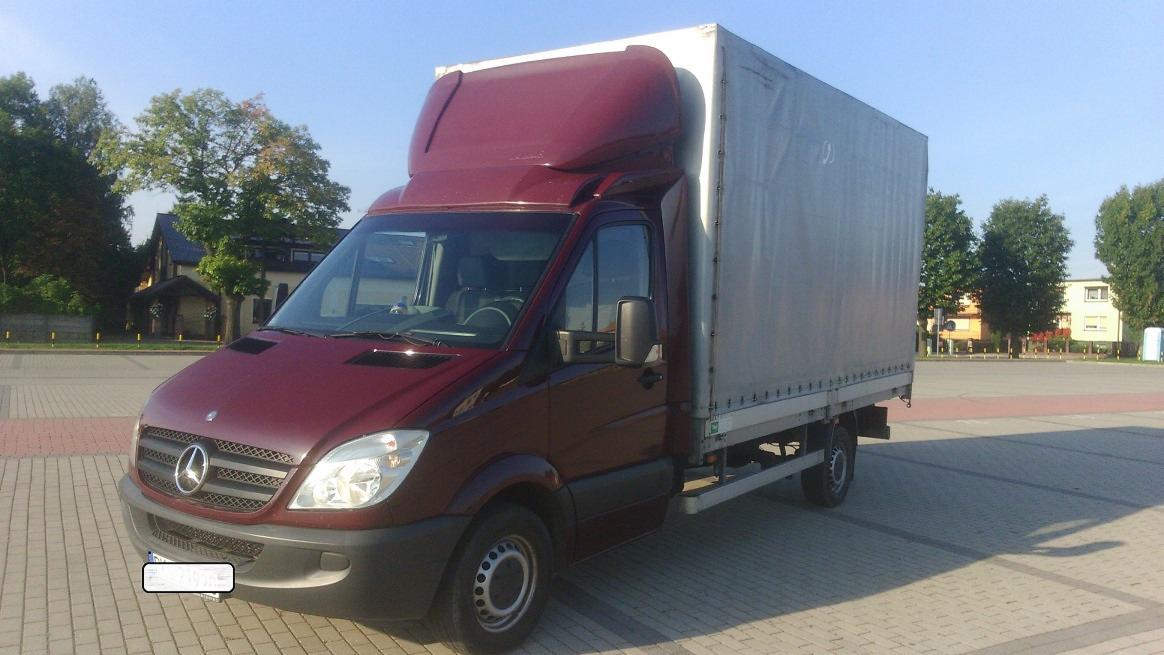 Czerwony bus Mercedes-Benz z plandeką na parkingu w Śmiglu, widok z przodu i boku, z budynkami w tle.