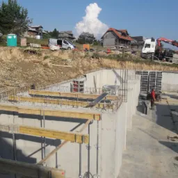 Betonowy fundament domu w trakcie budowy, widoczne zbrojenia, szalunki, drewniane podpory, w tle krajobraz wiejski z domami i dźwigiem.