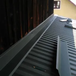 Fragment dachu z blachy trapezowej w trakcie montażu obróbek blacharskich, widoczny ołówek stolarski na powierzchni dachu i fragment drewnianej ściany szczytowej budynku w tle.