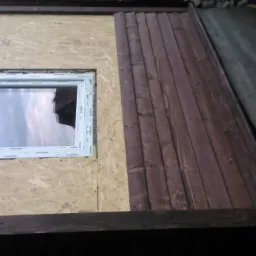 Fragment ściany zewnętrznej w trakcie budowy: okno PCV osadzone w płycie OSB, obok ciemne deski elewacyjne, widoczny fundament i poziomica.