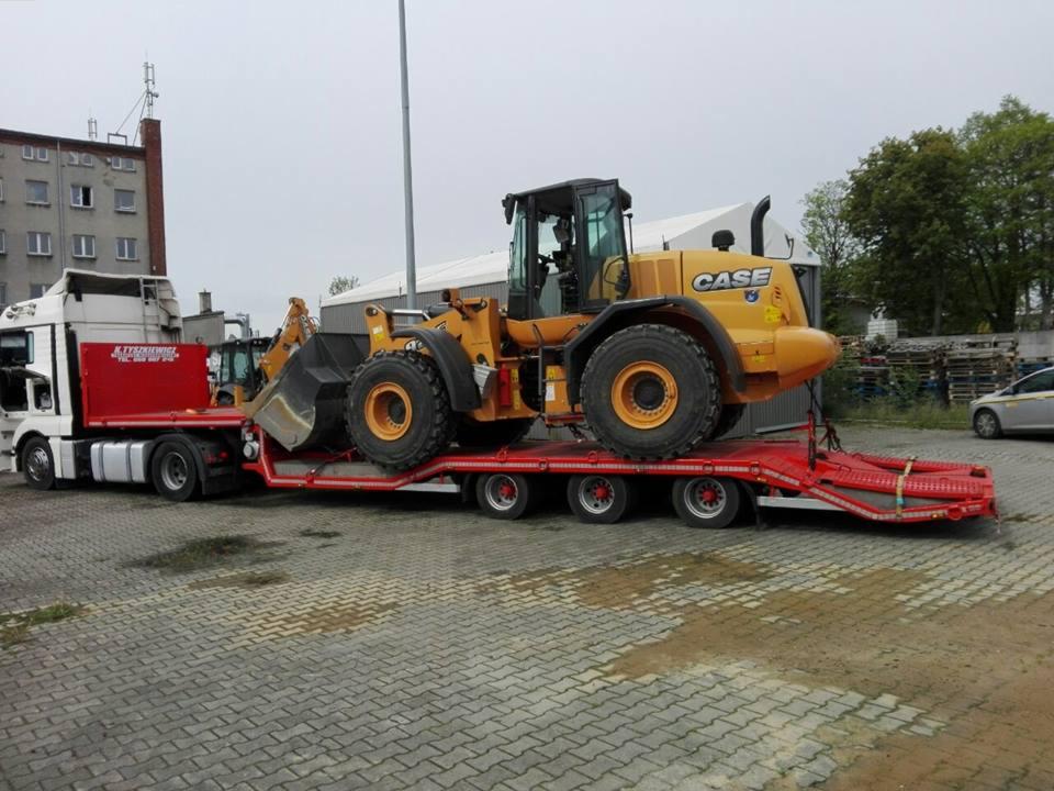 Żółta ładowarka CASE transportowana na czerwonej niskopodwoziowej naczepie ciągniętej przez białą ciężarówkę z czerwoną zabudową, widok z boku na brukowanym placu.