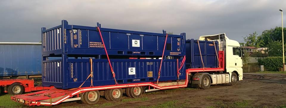 Ciężarówka z naczepą przewożąca dwa niebieskie kontenery zabezpieczone czerwonymi pasami transportowymi.