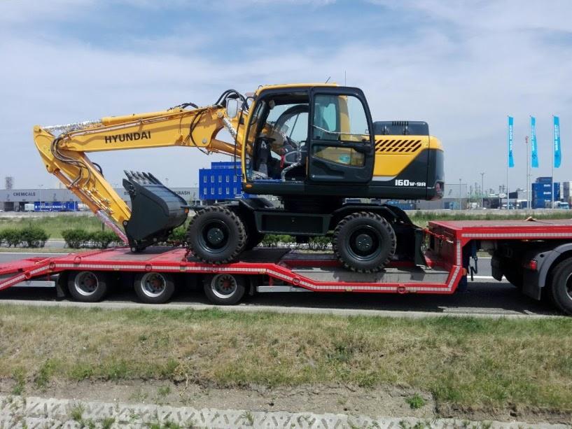 Żółta koparka Hyundai 160W-9A na czerwonej niskopodwoziowej platformie transportowej, widok z boku, w tle budynki przemysłowe.