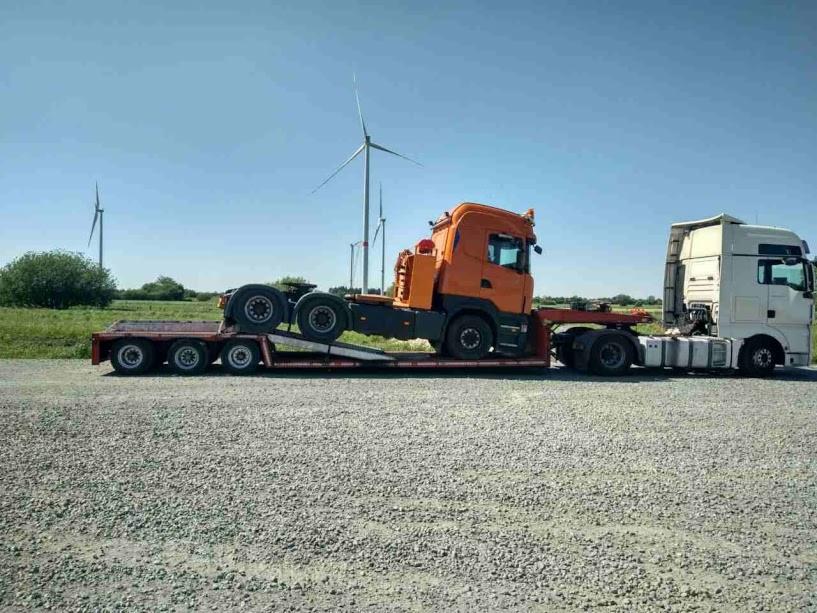 Pomarańczowy ciągnik siodłowy Scania przewożony na niskopodwoziowej platformie, z białym ciągnikiem z przodu, w tle turbiny wiatrowe.