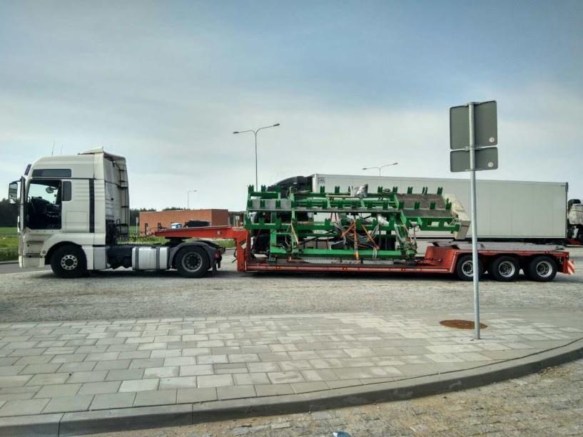 Ciężarówka z naczepą przewożąca zieloną konstrukcję stalową na tle pochmurnego nieba i brukowanej nawierzchni.