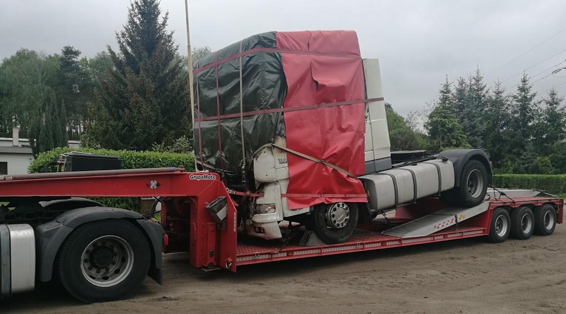 Ciężarówka zabezpieczona plandekami, transportowana na niskopodwoziowej platformie z rampą, widok z boku na tle zieleni.