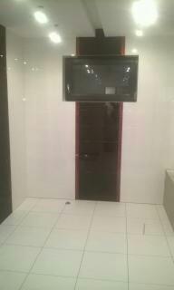 Biała, kwadratowa podłoga z płytek w pustym pomieszczeniu z białymi ścianami, na których wisi duży, czarny telewizor zamontowany na ciemnoczerwonej, pionowej płycie. Widoczne punktowe oświetlenie...