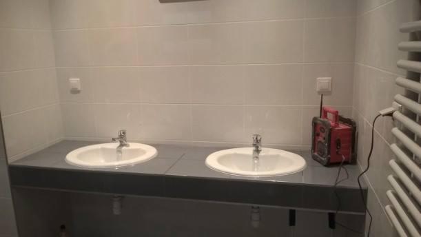 Dwie białe, okrągłe umywalki z chromowanymi bateriami na szarym blacie w łazience wyłożonej białymi, podłużnymi płytkami. Obok umywalki stoi czerwone radio budowlane podłączone do grzejnika.