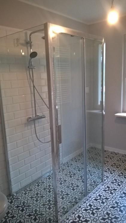 Nowoczesna kabina prysznicowa z przesuwnymi drzwiami, białe kafelki typu metro na ścianie, czarno-biała wzorzysta podłoga, widoczny grzejnik drabinkowy w tle.