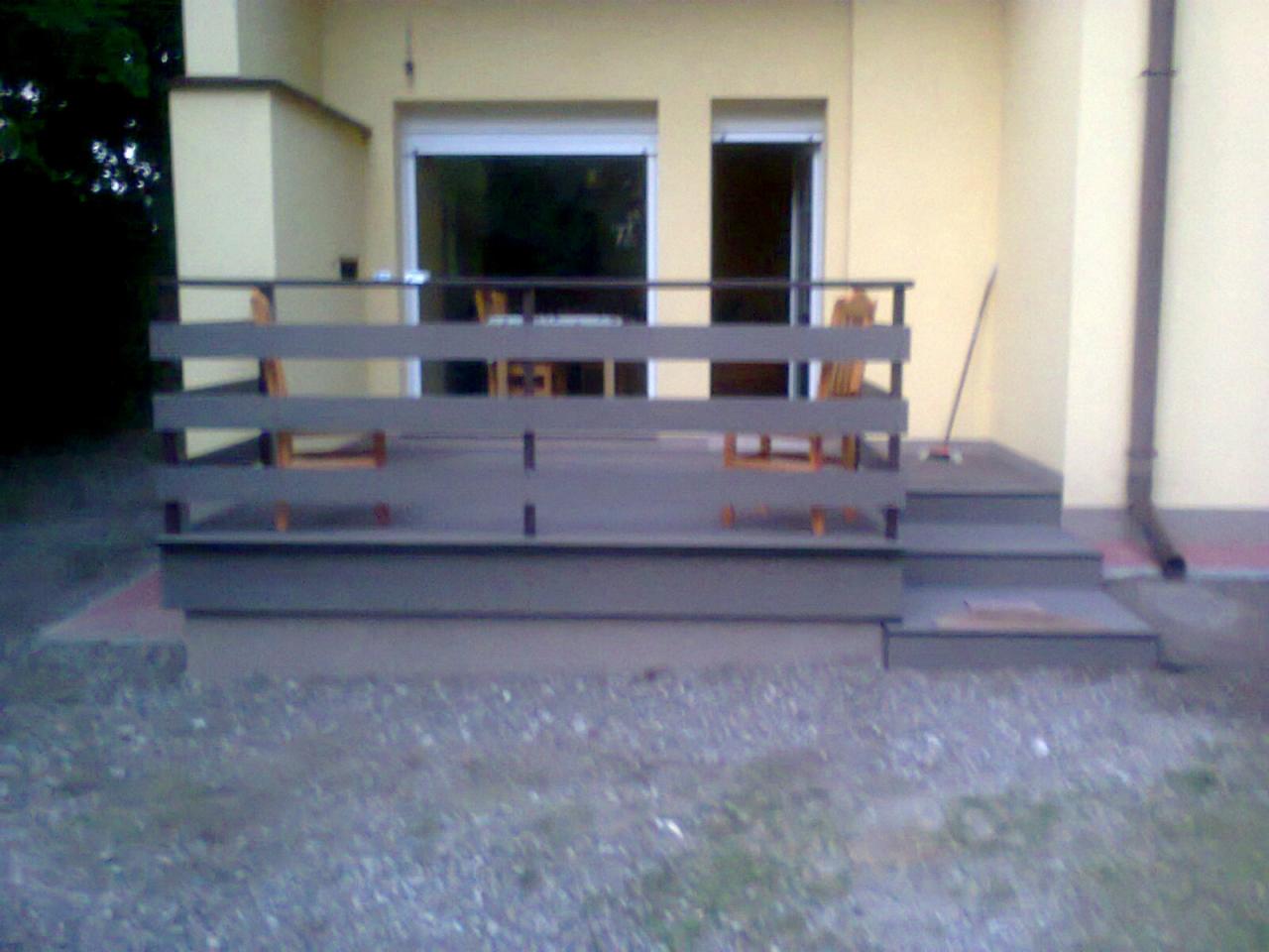 Nowo wybudowany szary taras z drewnianą balustradą i schodami przy żółtym domu, widok z niskiej perspektywy.