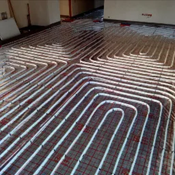 Białe rury ogrzewania podłogowego ułożone w pętle na czerwonej siatce montażowej, widoczne elementy instalacji w surowym stanie.