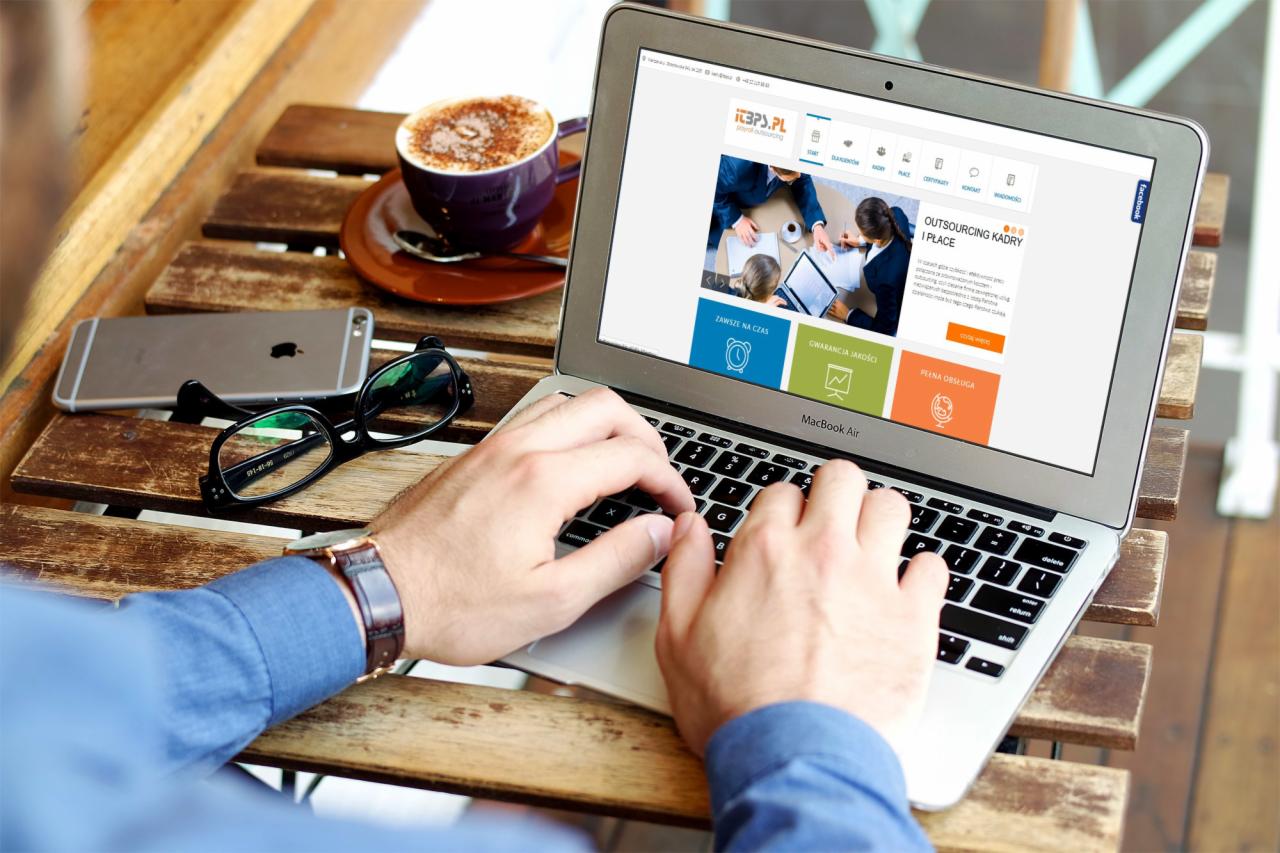 Mężczyzna pisze na laptopie MacBook Air na drewnianym stole z filiżanką cappuccino, okularami i smartfonem; na ekranie laptopa strona internetowa firmy oferującej outsourcing kadr i płac.