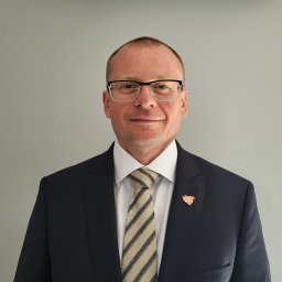 CONCORDIA FINANSE SPÓŁKA Z OGRANICZONĄ ODPOWIEDZIALNOŚCIĄ - Portret mężczyzny w garniturze z przypinką, uśmiechniętego, w okularach, na jasnoszarym tle. Profesjonalny wizerunek doradcy finansowego.