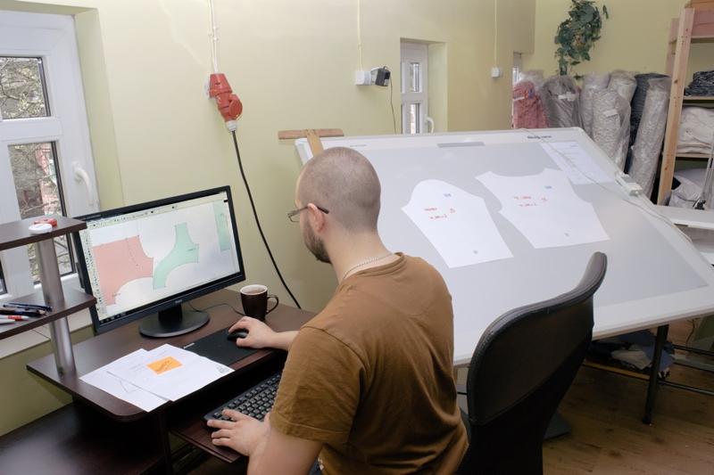 Projektant odzieży przy biurku, pracuje nad cyfrowym wzorem ubrania na monitorze komputera, obok kubek z kawą, na stole roboczym podświetlane szablony odzieży.