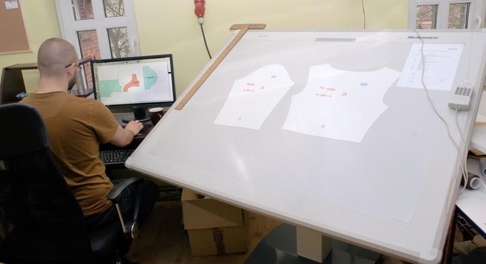 Projektant odzieży pracuje przy komputerze, obok duży stół kreślarski z szablonami wykrojów i drewnianą linijką typu litera T.