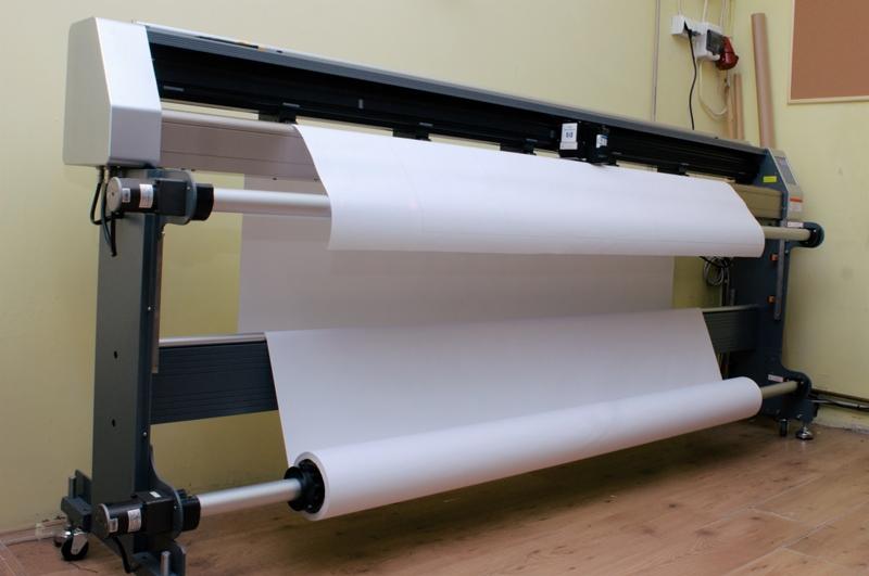 Plotter tnący z nawiniętym białym papierem, widoczne rolki i mechanizm przesuwu, oświetlenie studyjne.